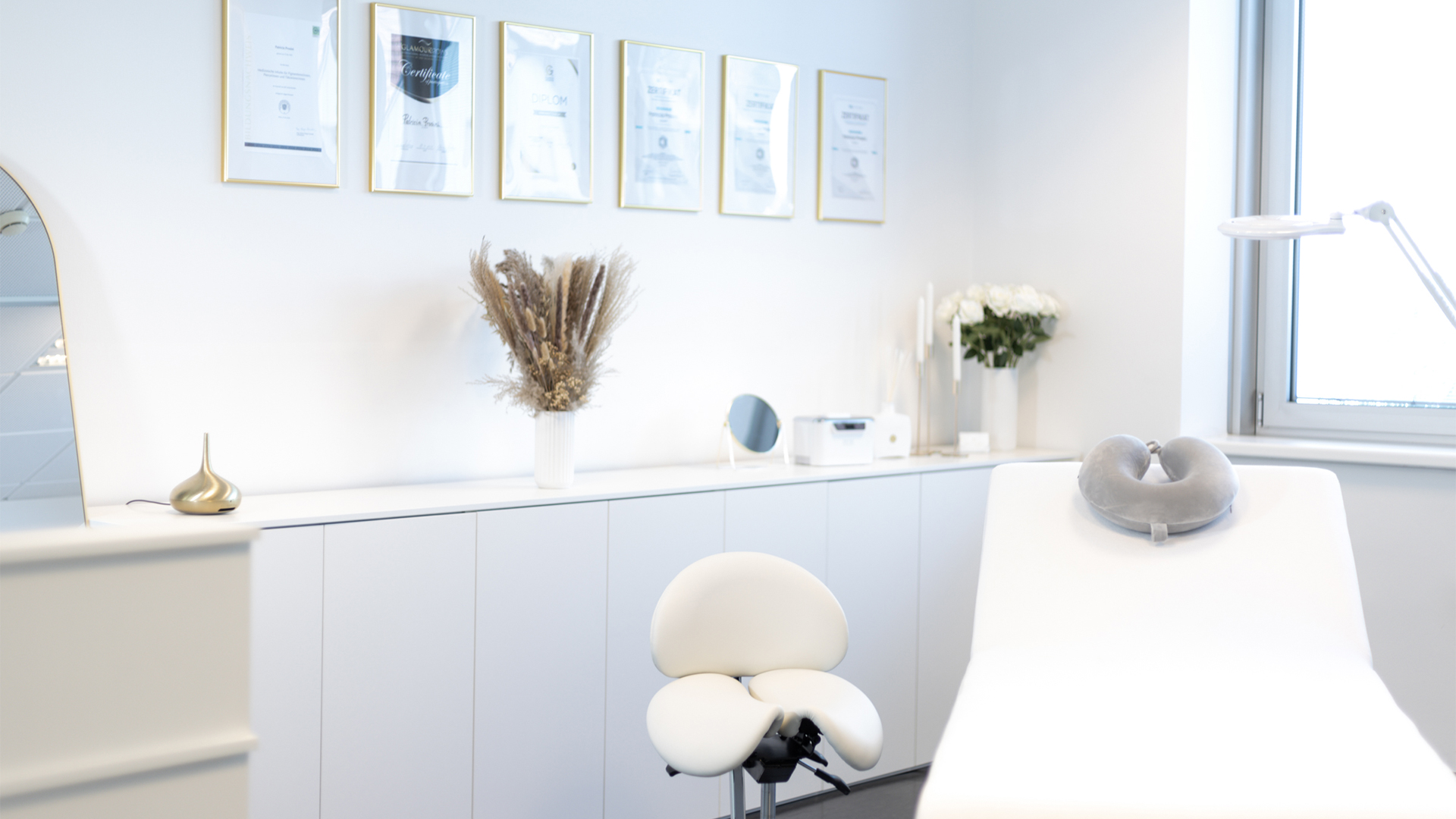 Foto von BEAUTY DREAMS Studio. Im Vordergrund die Behandlungsliege, der Stuhl, eine Lampe. Im Hintergrund Zertifikate in Rahmen an der Wand, ein SIdeboard und Dekoration